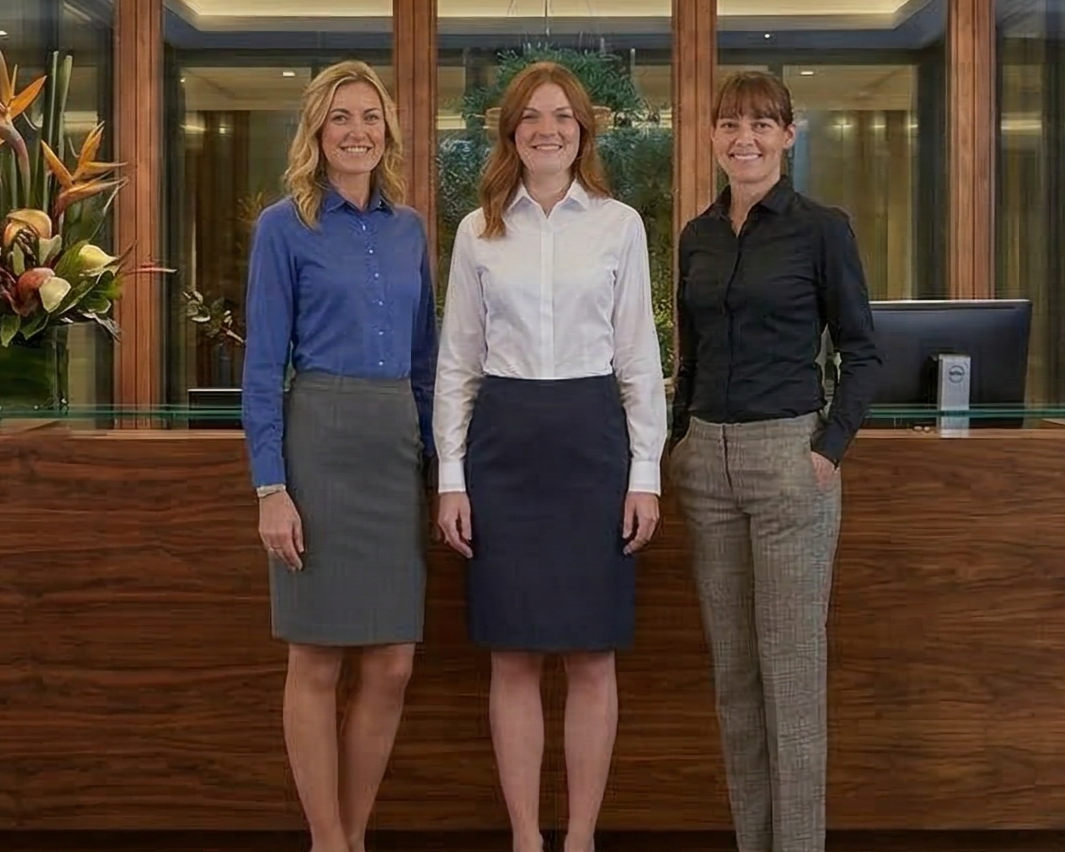 Three women standing in a modern lobby with wooden floors and glass walls.